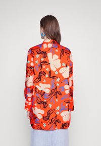 Blouse florale rouge à coupe ample, avec de grandes fleurs et feuilles blanches, bleues et vertes encadrées de noir. Manches longues et col.
