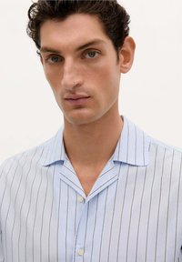 Camisa a rayas azul claro con una textura suave, cuello corto y dos botones. Presenta rayas verticales en azul más oscuro.