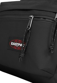 Schwarzer Stoff-Rucksack mit Reißverschlusstasche, ausgestattet mit einem Eastpak-Logo-Patch, strukturierter Oberfläche und geflochtenen Zugbändern.