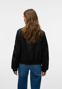 Veste bomber noire avec des poignets et un ourlet froncés, dotée d'une texture lisse et de manches rembourrées. Portée avec un jean bleu taille haute.