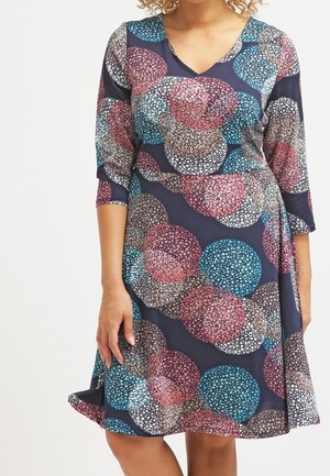 Vrouw draagt een knielange marineblauwe jurk met driekwartmouwen en overlappende ronde mozaïekpatronen in roze, blauw, wit en beige.