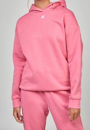 Personne portant un sweat à capuche rose et un pantalon assorti avec un petit logo blanc sur la poitrine, debout devant un fond clair.