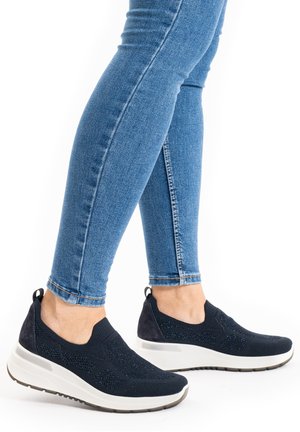 Beine in blauen engen Jeans, die schwarze Slip-On-Sportschuhe mit weißen dicken Sohlen tragen, stehend vor weißem Hintergrund.