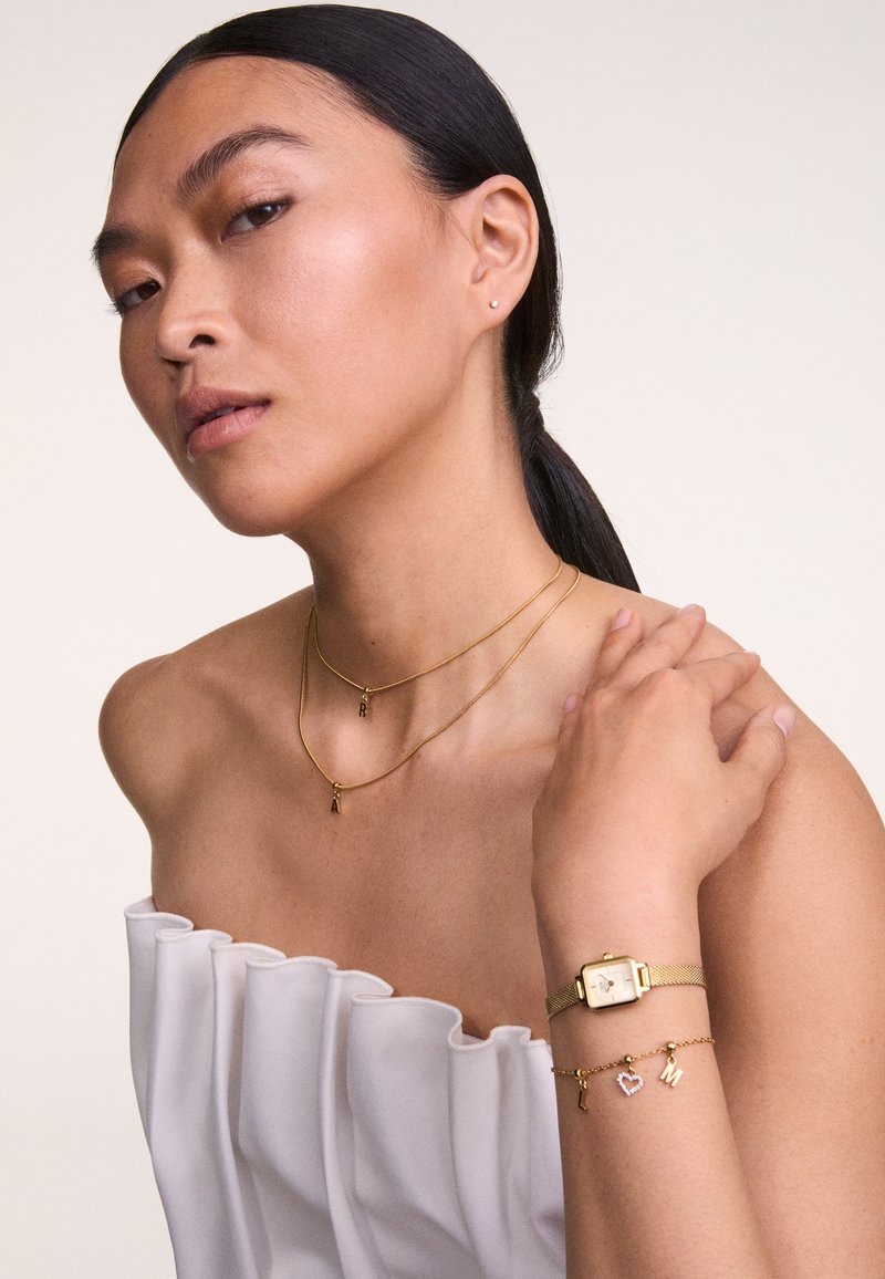 Goldene Halsketten mit Anhängern, goldene Uhr mit Mesh-Armband, Armband mit Charms, Modell trägt ein weißes, rüschendes Oberteil, neutraler Hintergrund.