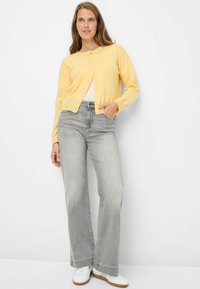 Cardigan giallo con scollo rotondo e chiusura con bottoni, abbinato a jeans grigi chiaro a gamba larga e sneakers bianche. Tessuto liscio, vestibilità casual.