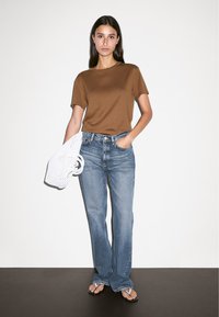 Brown, short-sleeve t-shirt paired with light blue, straight-leg jeans. Holds a white, woven bag and wears brown flip-flops. Minimal background.