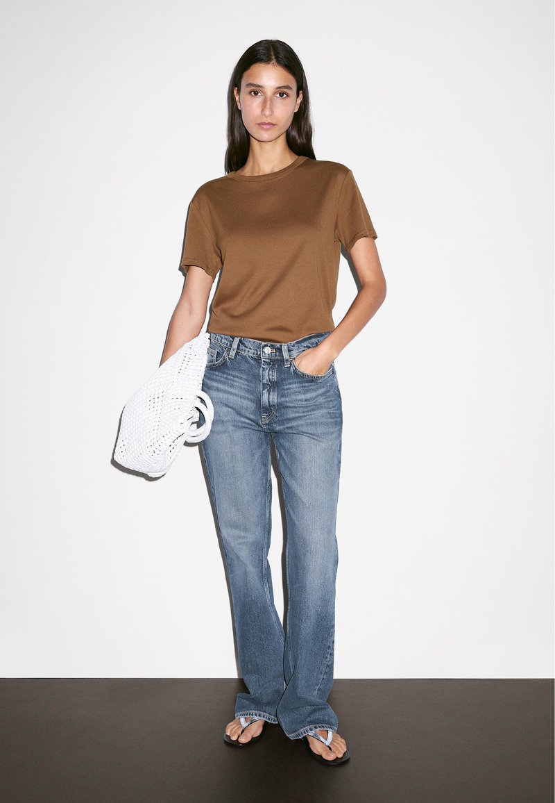 Brown, short-sleeve t-shirt paired with light blue, straight-leg jeans. Holds a white, woven bag and wears brown flip-flops. Minimal background.