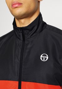 Chaqueta negra con cremallera y cuello alto, con una franja roja horizontal y un logo circular en el lado izquierdo del pecho. Tejido texturizado.