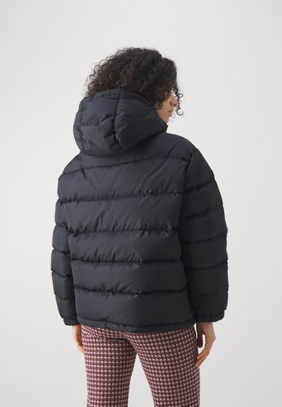 Crna puffer jakna s kapuljačom, s horizontalno quilted dijelovima i glatkom teksturom. Nosena preko uzorčatih tajica, prikazivanje stražnjeg pogleda.