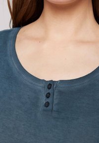 Krátký rukáv, tmavě modrá bavlněná košile s kulatým výstřihem a třemi černými knoflíky na légě. Měkká textura a přiléhavý střih.