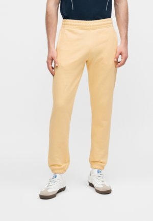 Homme portant un pantalon de jogging beige et des baskets blanches, debout sur un fond uni clair.