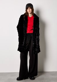 Czarny płaszcz z sztucznego futra z czerwonym prążkowanym topem. Czarne szerokie spodnie i masywne czarne platformy. Czarna czapka podkreśla wygląd.