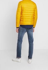 Gul puffjakke med vandret syninger, marineblå manchetter, parret med blå jeans og hvide sneakers. Synlige detaljer inkluderer baglommer.