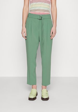 Personne portant un pantalon vert taille haute avec une ceinture, un top sans manches à carreaux multicolore et des mocassins vert clair, debout devant un fond uni.