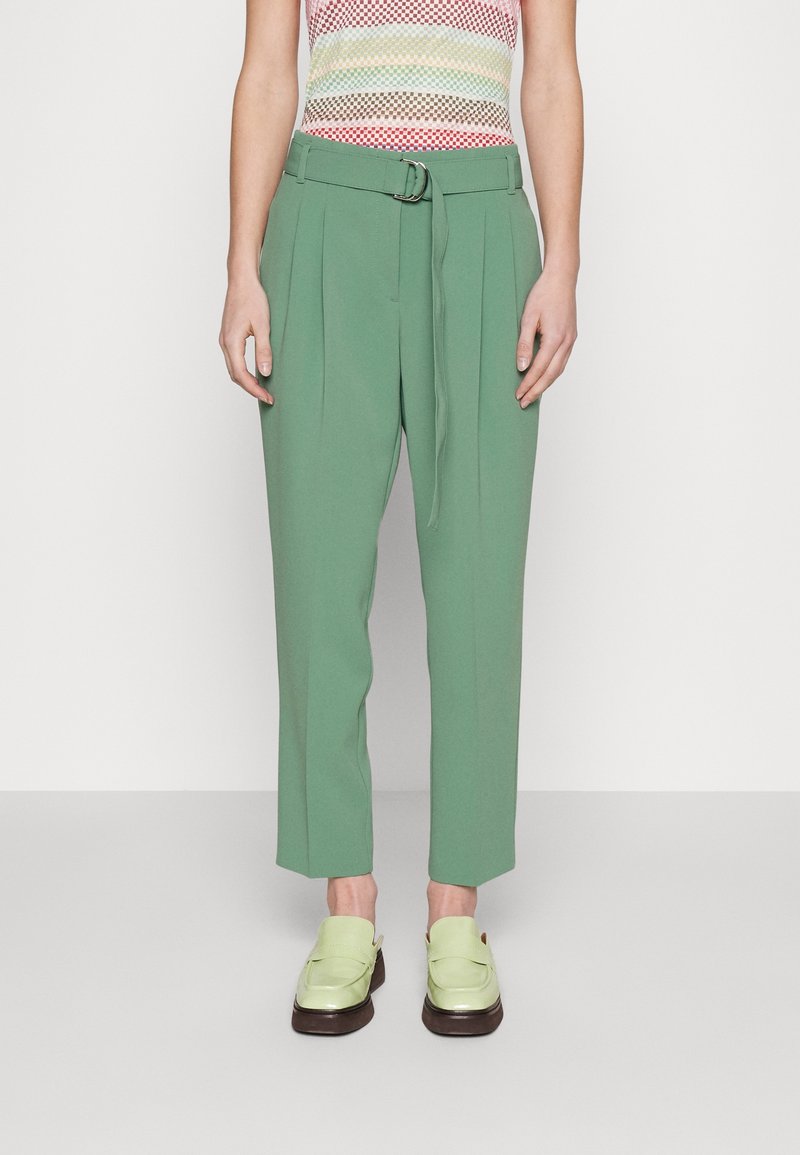 Personne portant un pantalon vert taille haute avec une ceinture, un top sans manches à carreaux multicolore et des mocassins vert clair, debout devant un fond uni.