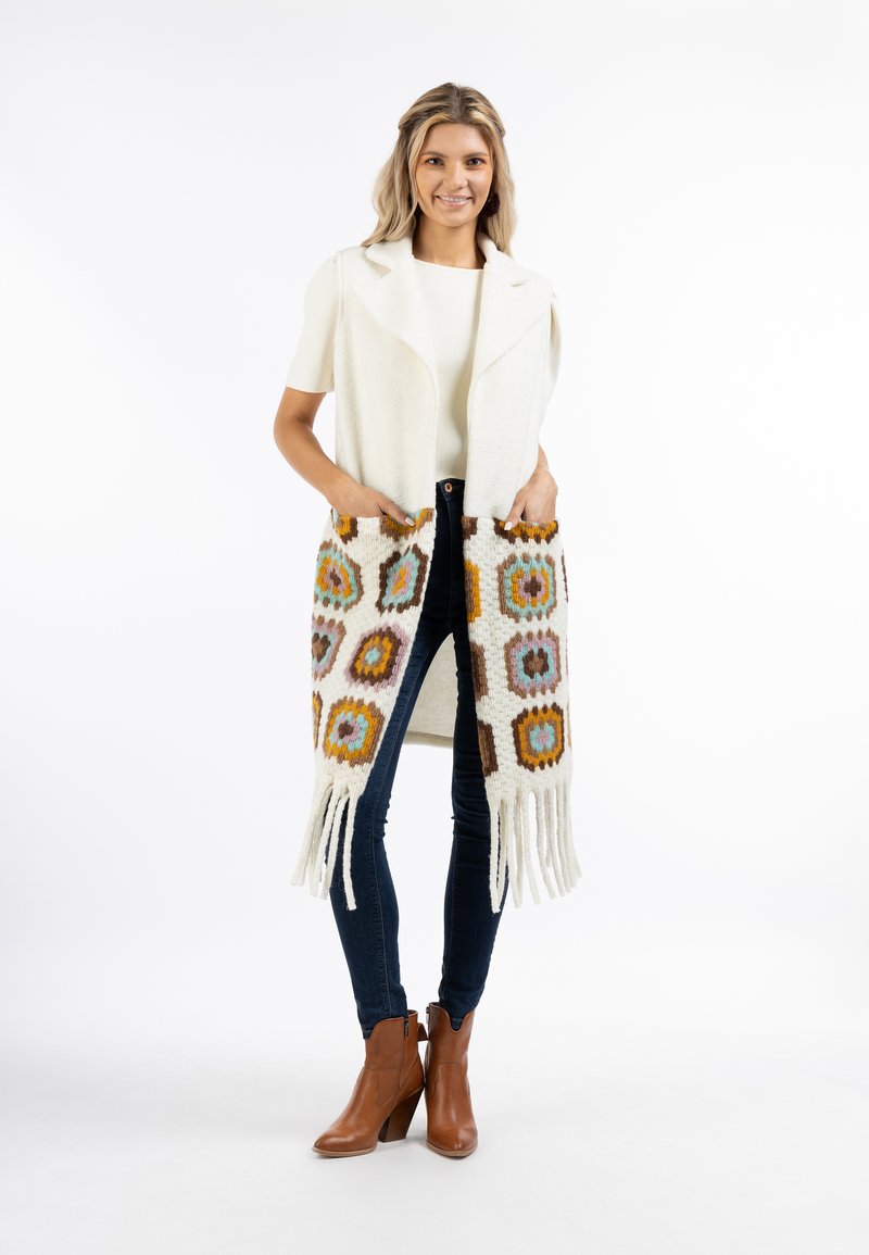 Strickweste in Creme mit gemusterten Taschen, die hexagonale Formen in Orange, Blau und Braun zeigen; fransige Kanten; getragen über einem hellen Oberteil und Jeans.