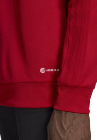 Felpa sportiva rossa con polsini e pannelli laterali a coste, realizzata in tessuto testurizzato. Logo 'AEROREADY' riportato in basso a destra.