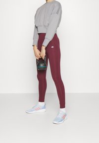 Sweatshirt grigio corto, leggings bordeaux a vita alta e sneakers azzurri chiari con dettagli rosa. Tiene in mano un kettlebell nero.