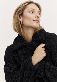 Palton negru din fleece cu guler lat, textură moale și croială lejeră. Cercei aurii adaugă un detaliu de contrast. Modelul ține paltonul de guler.