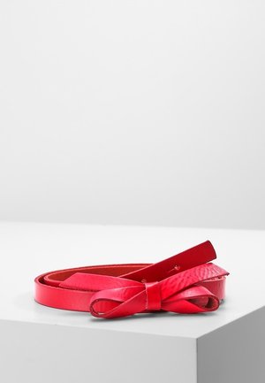 Ceinture en cuir rouge avec un détail de nœud bouclé, enroulée et reposant sur une surface blanche sur un fond gris clair uni.