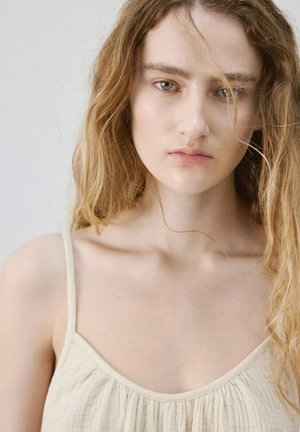 Joven mujer con cabello largo y ondulado rubio, vestida con una blusa sin mangas de color crema, sobre un fondo claro y sencillo.