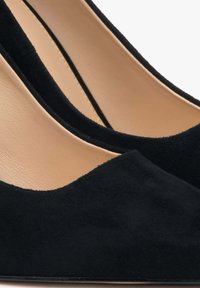 Černé semišové lodičky na vysokém podpatku s špičatou špičkou a hladkým, béžovým vnitřním podšíváním. Podpatek je štíhlý a úzký, což zvýrazňuje elegantní siluetu.