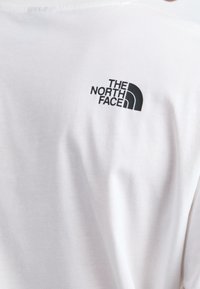 Weißes Baumwoll-T-Shirt mit einem kleinen schwarzen Logo von "The North Face" in geschwungener Schrift auf dem oberen Rücken, in der Nähe des Halsauschnitts.