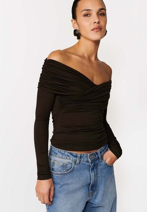 Kobieta ubrana w czarną bluzkę z marszczeniami i odsłoniętymi ramionami oraz długimi rękawami, w niebieskie jeansy z wysokim stanem, pozująca z jedną ręką w kieszeni na białym tle.