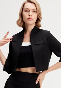 Veste noire courte avec un col haut, des épaules légèrement structurées et un détail de poche. Portée sur un haut noir ajusté. Texture de tissu lisse.