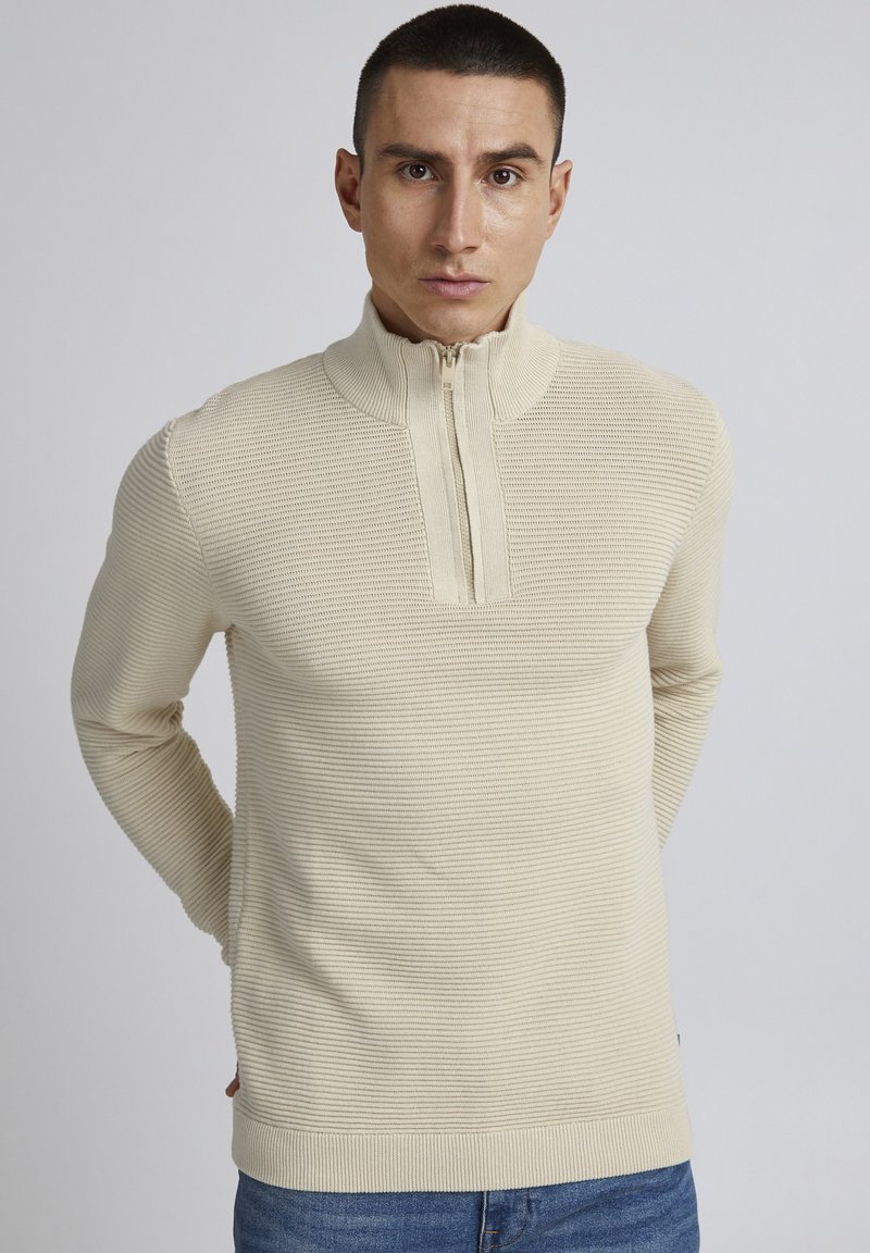 Beige gerippter Pullover mit Reißverschluss, hohem Kragen und langen Ärmeln. Glatte Textur und figurbetontes Design mit einem subtilen Glanz.