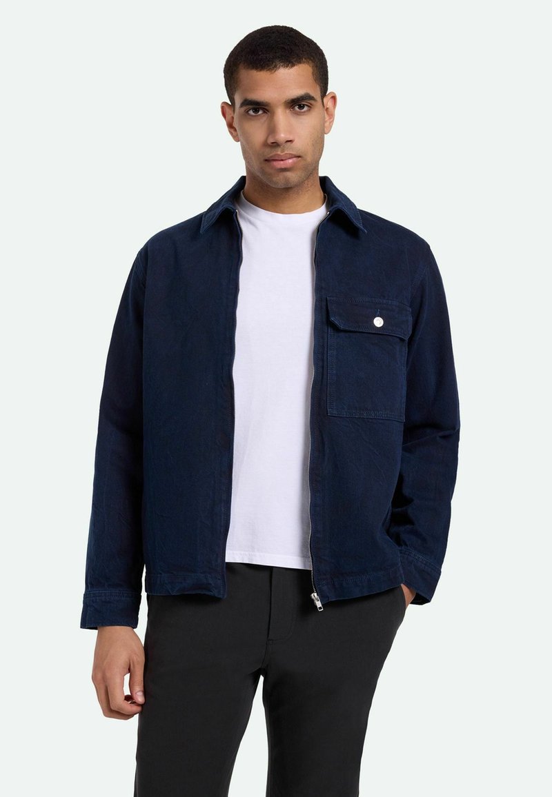 Granatowa kurtka z trwałego materiału, z zamkiem błyskawicznym z przodu, spiczastym kołnierzem oraz jedną kieszenią na piersi z okrągłym detalem guzikowym.