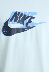 Světle modré bavlněné tričko s velkým černým a modrým grafickým logem Nike. Zaoblený výstřih a krátké rukávy. Hladká textura.