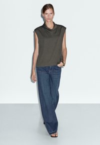 Oliwkowy bezrękawnik z drapowanym dekoltem, w zestawieniu z luźnymi ciemnoniebieskimi dżinsami. Modelka stoi na jasnym tle.