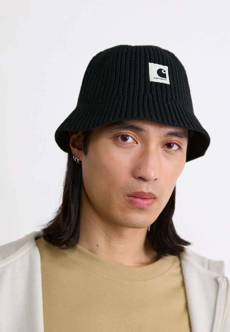 Čierny pletený klobúk v tvare vedra s rebrovanou textúrou a štvorcovou nášivkou. Šilt je široký, čo ponúka ležérny a funkčný dizajn.