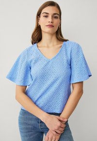 Kvinde iført en lyseblå bluse i øjletstof med flagrende ærmer og V-hals, kombineret med jeans i mellemblå vask mod en ensfarvet baggrund.