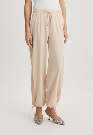 Pantalons beiges fuselés avec une taille élastique, un cordon de serrage et des panneaux latéraux en mesh ; associés à des chaussures à talons hauts à motifs.