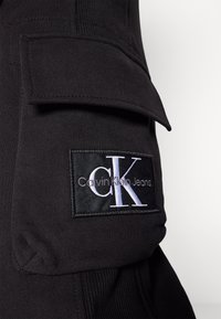 Svarta bomullscargobyxor med en sidficka och en etikett med "Calvin Klein Jeans" och initialerna "CK" i vitt.