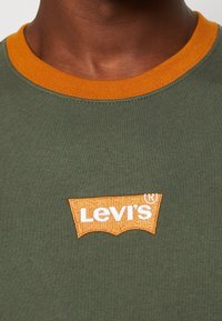 T-shirt verde oliva con scollatura a coste arancione. Presenta il logo ricamato di Levi's in bianco su uno sfondo arancione a contrasto.