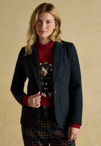 Tmavomodrý herringbone blejzer s dvoma prednými vreckami, skombinovaný s červeným sveterom s motívom psa a károvanými nohavicami v zelenej, červenej a žltej farbe.