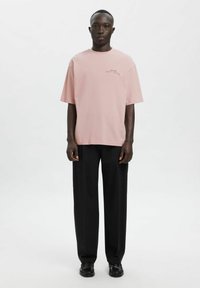 Jasnoróżowa, oversize'owa koszulka z krótkimi rękawami i detalem z logo, zestawiona z czarnymi spodniami o szerokich nogawkach oraz czarnymi butami. Prosty, czysty design.