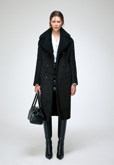 Femme portant un long manteau noir avec un grand col, des bottes noires montant jusqu'aux genoux, tenant un sac à main noir, debout sur un fond clair uni.