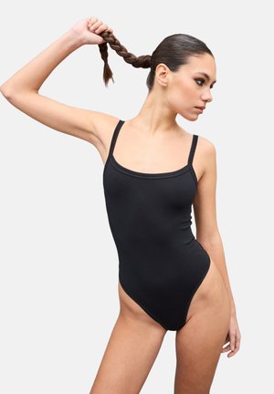 Mujer joven con un body negro sin mangas, sosteniendo su coleta trenzada hacia arriba y mirando hacia un lado, sobre un fondo claro y liso.