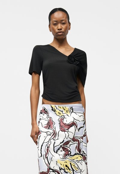 Γυναίκα που φοράει μαύρο ασύμμετρο τοπ με λεπτομέρεια από υφασμάτινο λουλούδι και λευκή φούστα με απεικονίσεις αγγέλων σε κόκκινο, κίτρινο και μαύρο χρώμα.