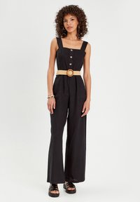 Black linen jumpsuit with wide legs, square neckline, front buttons, and a woven belt with a round buckle. Features two front pockets.