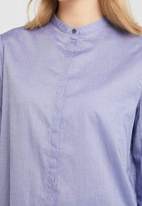 Camicia viola chiaro a maniche lunghe con colletto alla band. Presenta una leggera texture e un singolo bottone al collo, mostrando un design semplice.