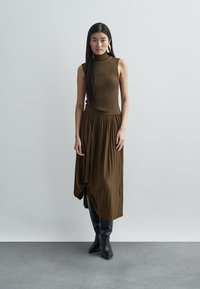 Brown ribbed long dress with a fitted bodice and pleated skirt, featuring a high neckline and a knotted detail at the hem. Black knee-high boots.