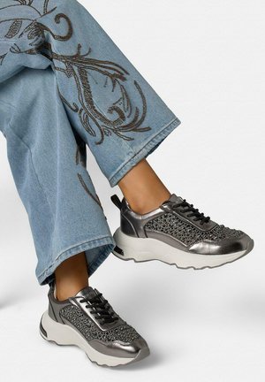 Persona con vaqueros azules con bordado y zapatillas negras metálicas con malla decorativa y suelas blancas gruesas, piernas cruzadas en el aire.