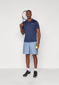 Tumesinise spordikuubiga lühikeste varrukatega särk, millele on kombineeritud helesinised ruudulised lühikesed püksid. Mustad spordijalatsid ja tennisereket, hoides palli.