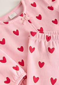 Rosa pyjamaset i två delar med en topp som har rund halsringning och hjärtmönster i rött med ljusgröna konturer, dekorerad med en rosett.