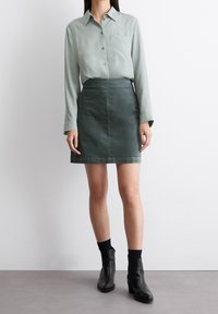 Camisa de manga larga verde claro con botones y un bolsillo en el pecho, combinada con una falda de mezclilla verde oscura. Botines negros y calcetines completan el look.